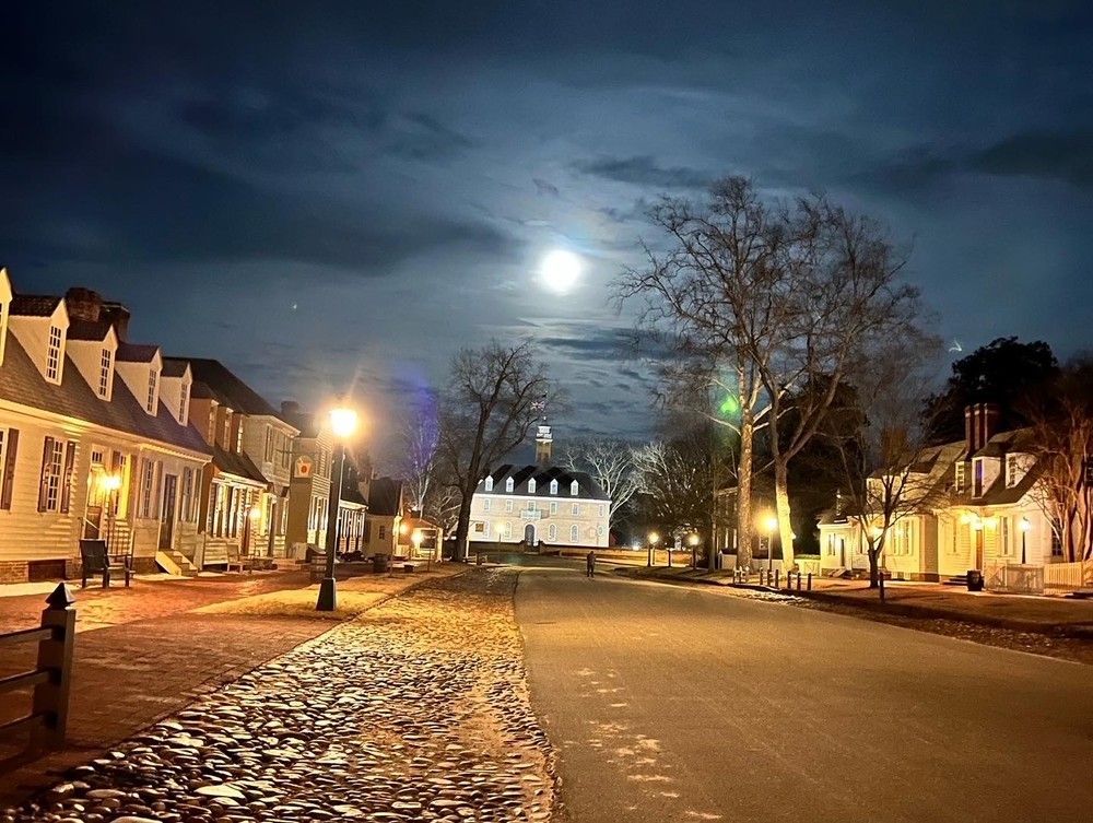 Cobblestone street at night, lined with colonial-style buildings and streetlights; full moon in the sky.