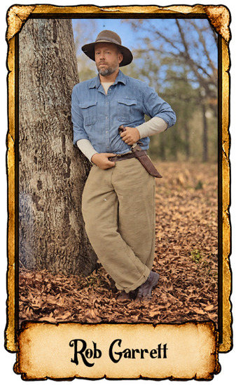 Man in gray hat and blue shirt leans against a tree in an autumn forest, holding a shotgun.