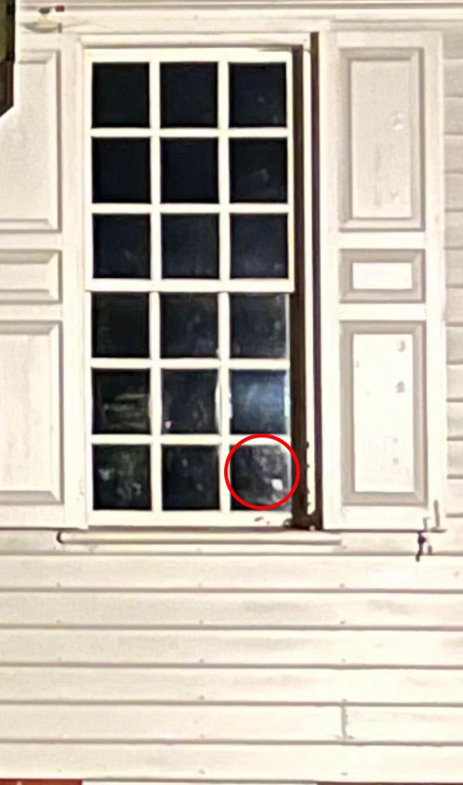 White-framed window with small panes, two closed shutters, on a white clapboard house. A dark figure is visible in the bottom right pane.