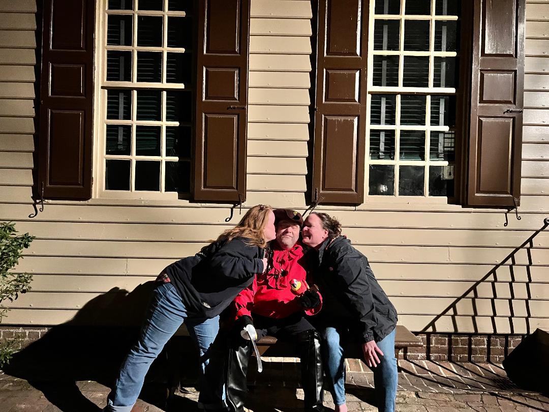 Two women kissing a person in a wheelchair in front of a building with brown shutters and windows.