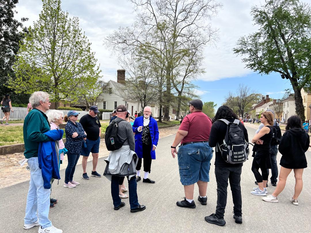 Group listening to a costumed person in blue coat on a street with historic buildings.