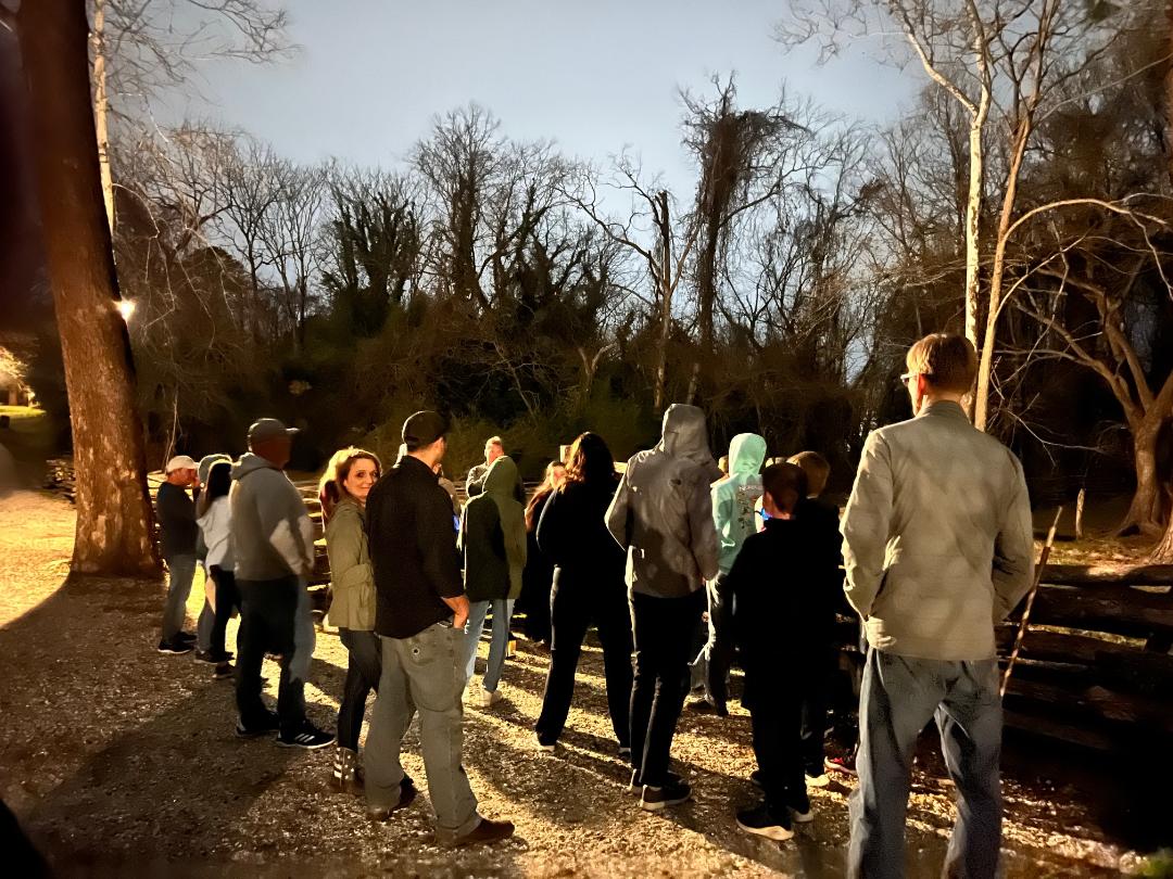A group of people standing outside at dusk, near trees. Some are looking towards the right.