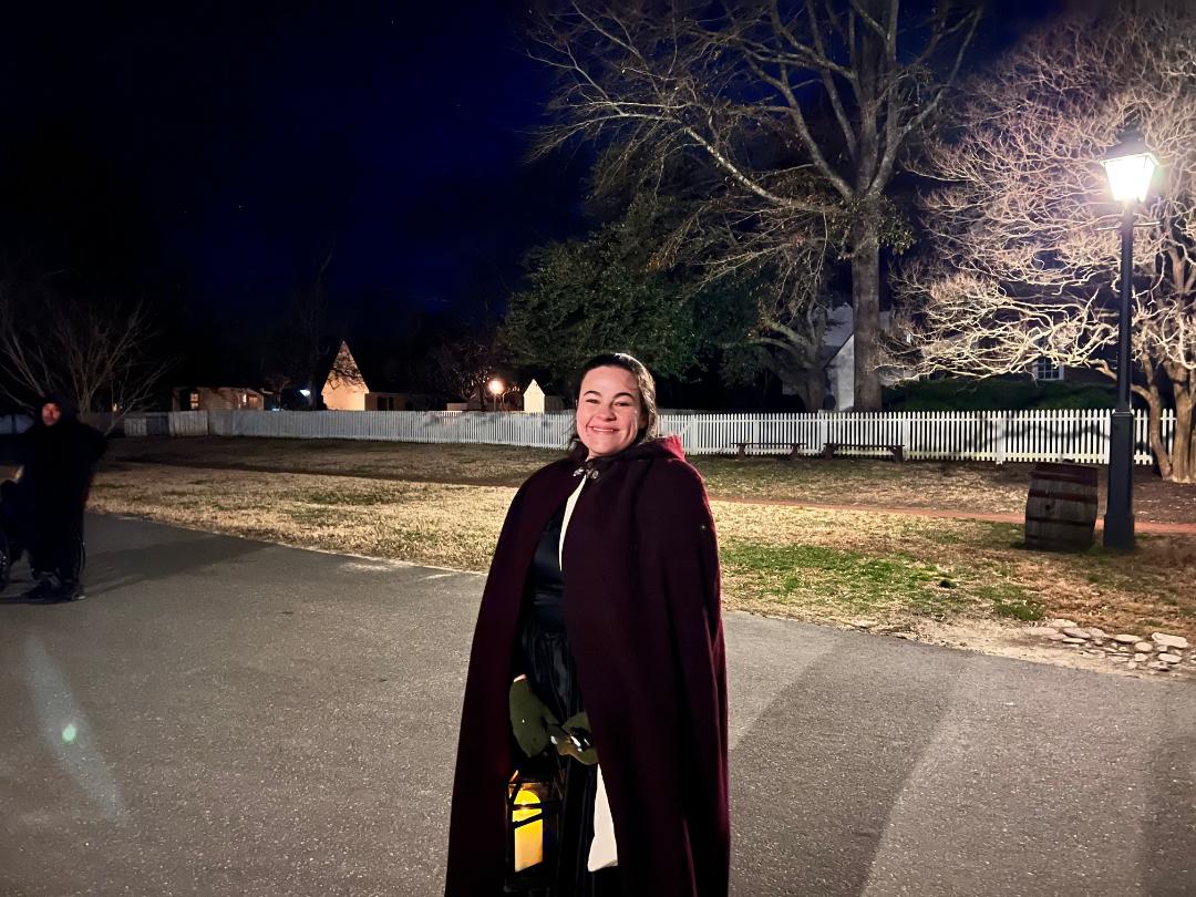 Woman in burgundy cape smiles outdoors at night, lamplit path, Colonial Williamsburg background.