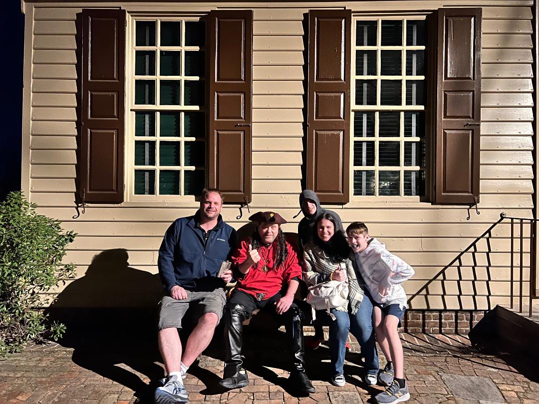 A group of five people seated on a bench in front of a building with brown shutters and windows.
