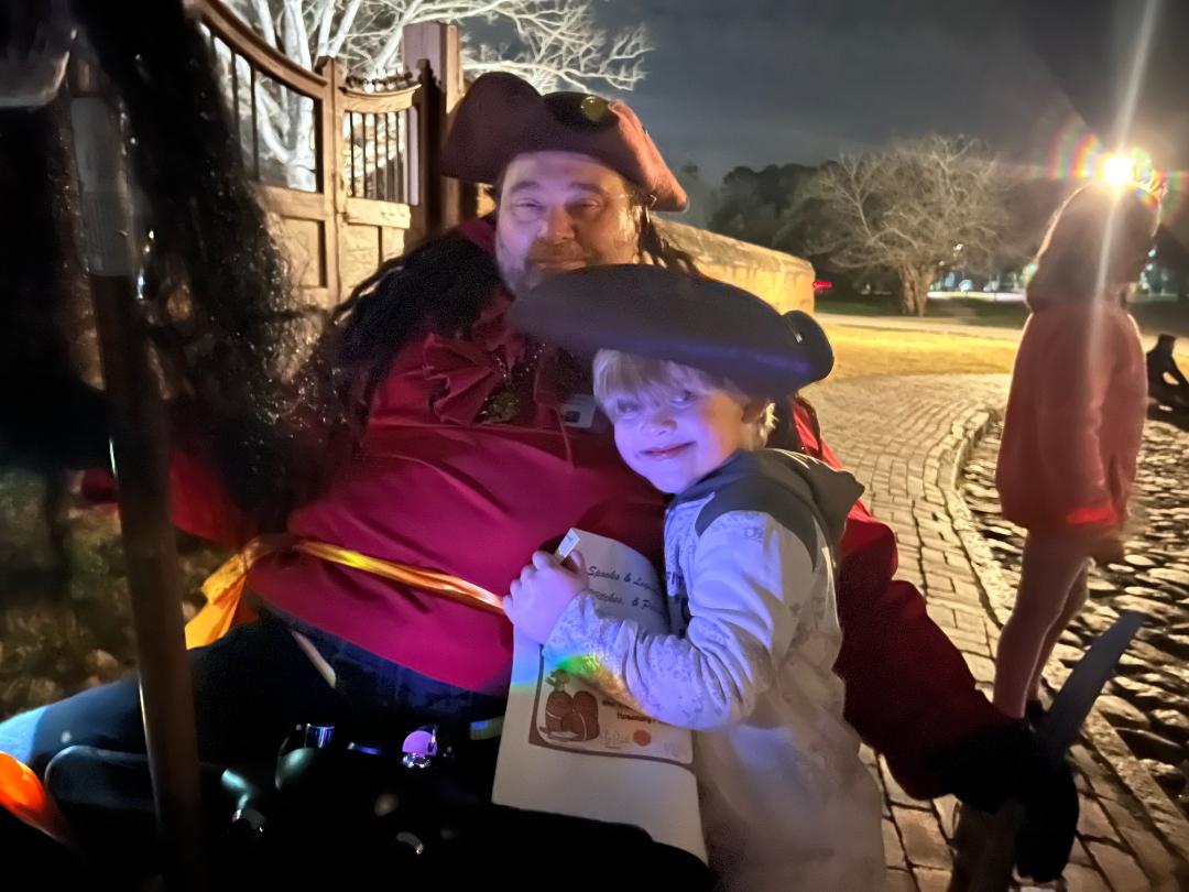Man in pirate costume and child smiling, both wearing hats, outdoors at night.