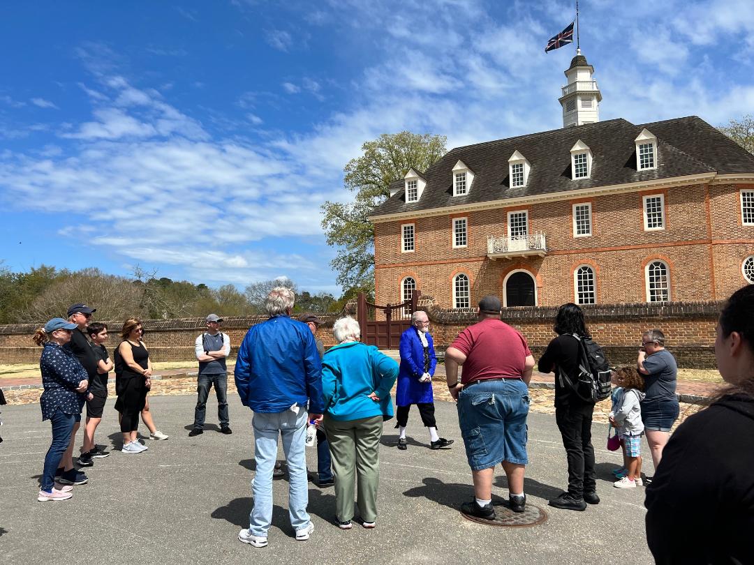 People gather for a tour in front of a brick building. A person in a blue coat speaks. Sunny day.