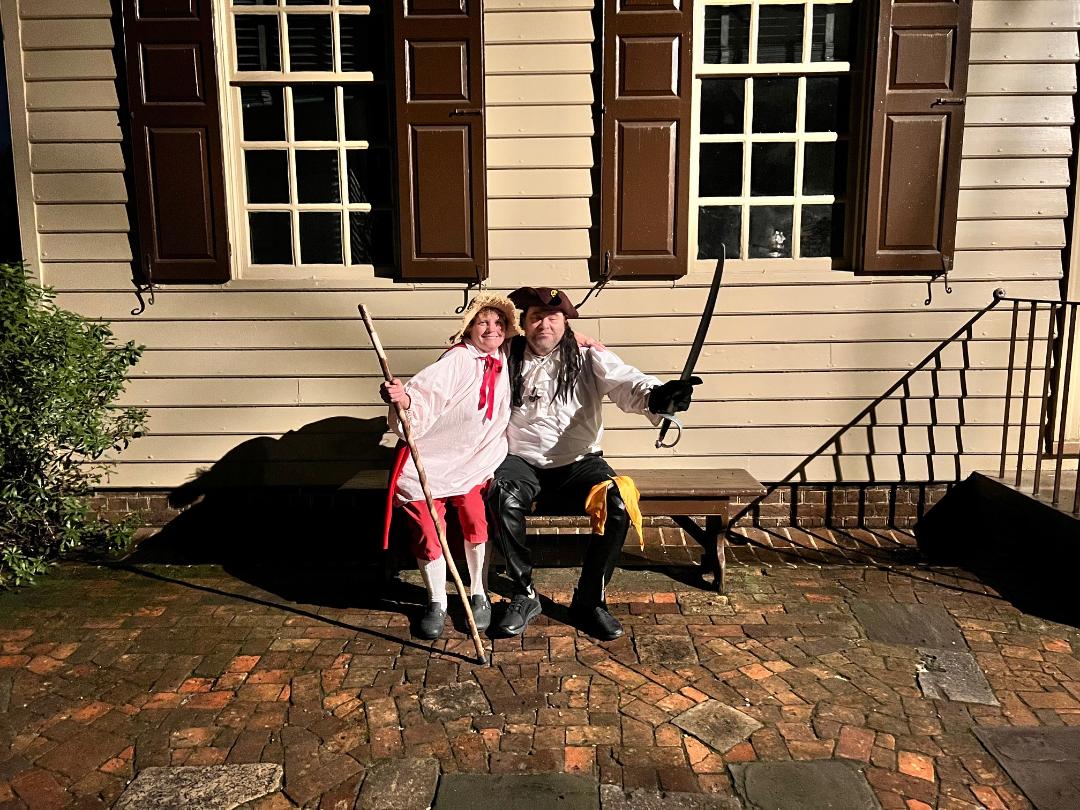 Two people in pirate costumes posing on a bench in front of a building with brown shutters.