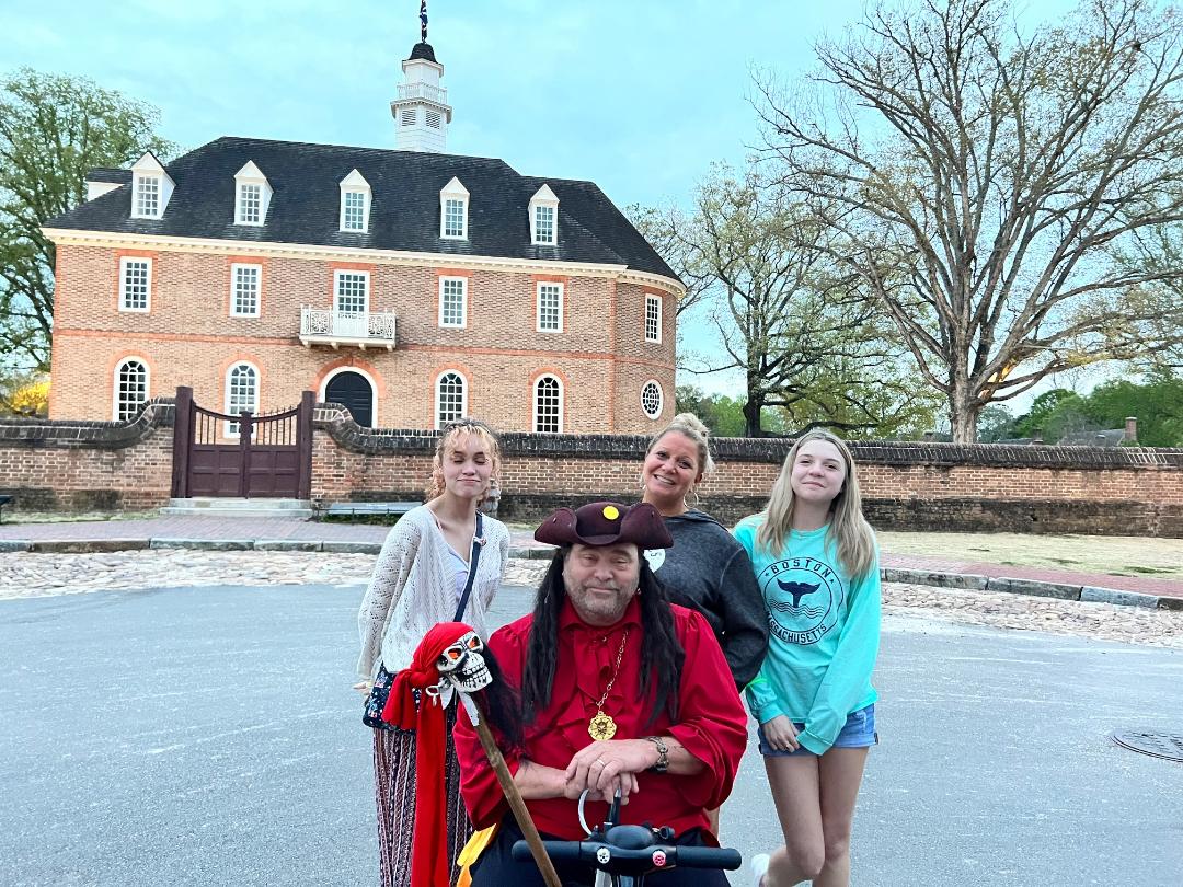 Four people pose in front of a colonial brick building with a spire. One holds a staff and wears a hat.