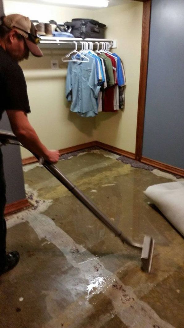 Person using a squeegee to clean water from a carpeted closet floor with hanging clothes nearby