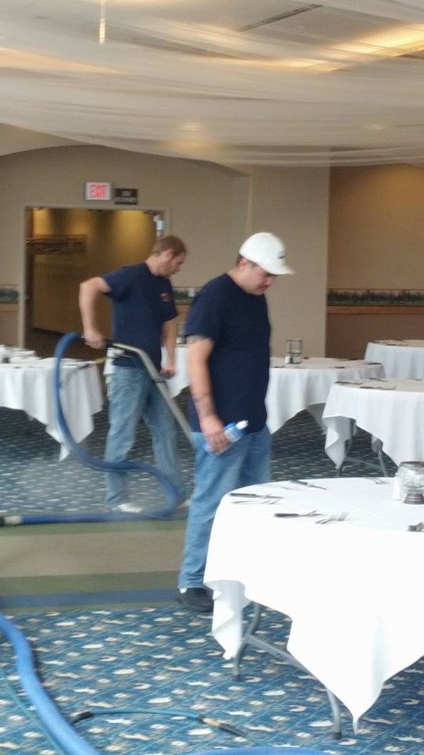 Two men moving furniture in a banquet room with round tables and blue carpet
