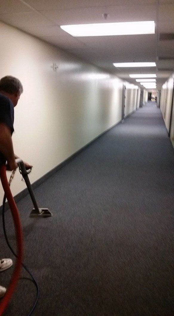 Person vacuuming a long, empty carpeted hallway with beige walls and ceiling lights