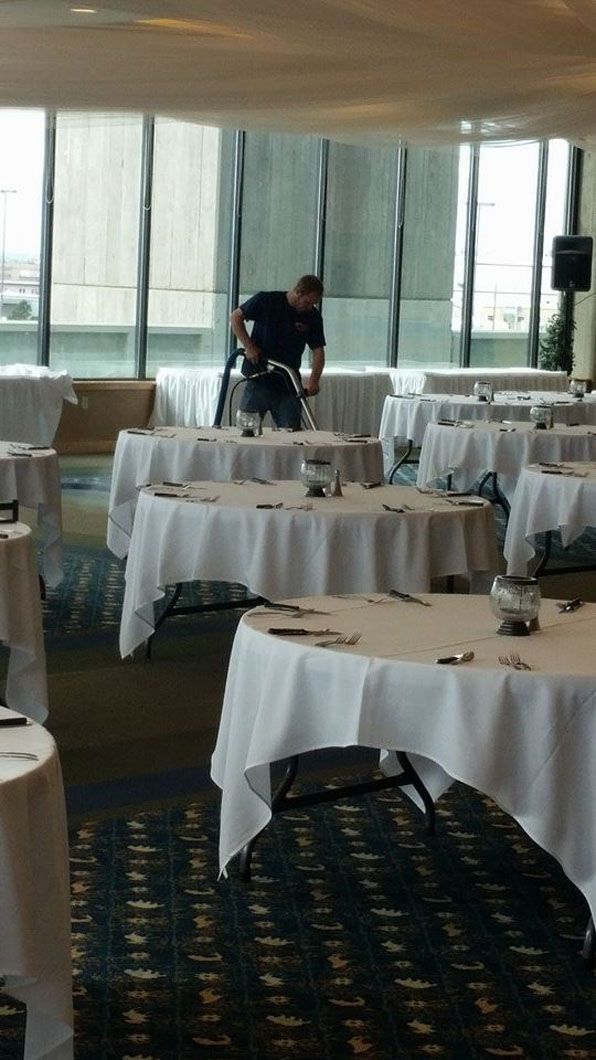 Empty banquet hall with round tables covered in white linens and two people setting up near windows.