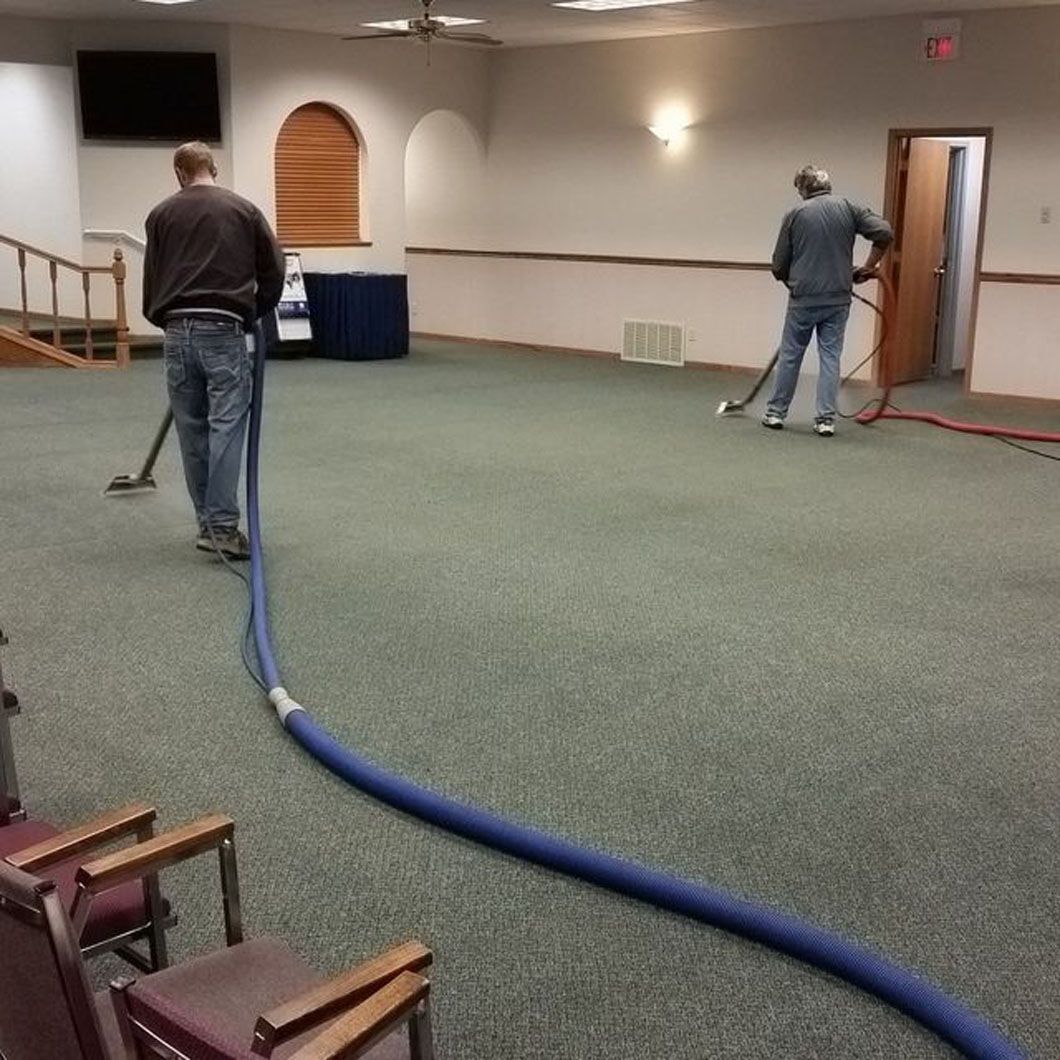 Two workers vacuuming carpet in a room, with blue hose stretched across the floor.
