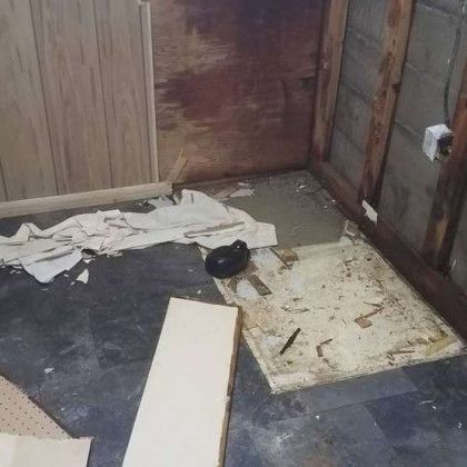 Dirty, damaged bathroom floor with torn paper, a black bowl, and broken tiles near wood-paneled walls