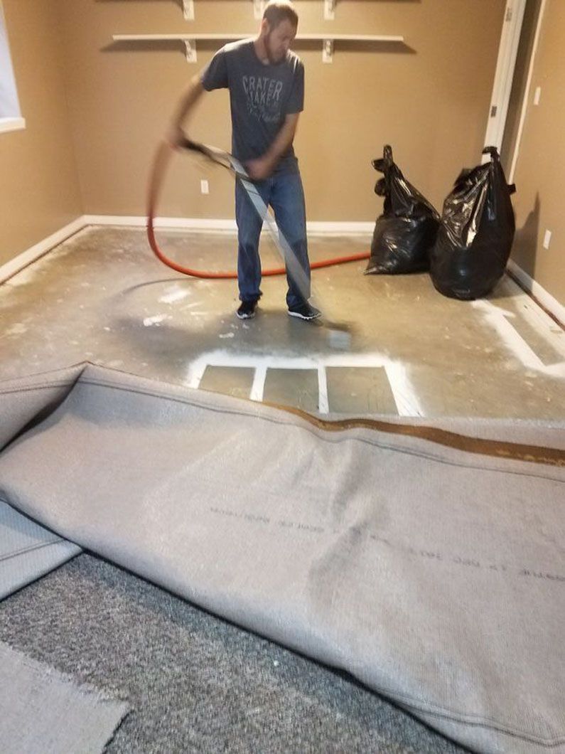 Man smoothing gray carpet padding in a room with tan walls and black trash bags nearby
