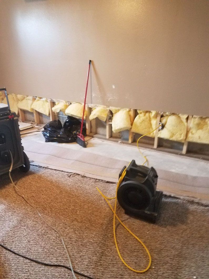 Basement wall with exposed insulation, a broom, and floor fans on a concrete floor.