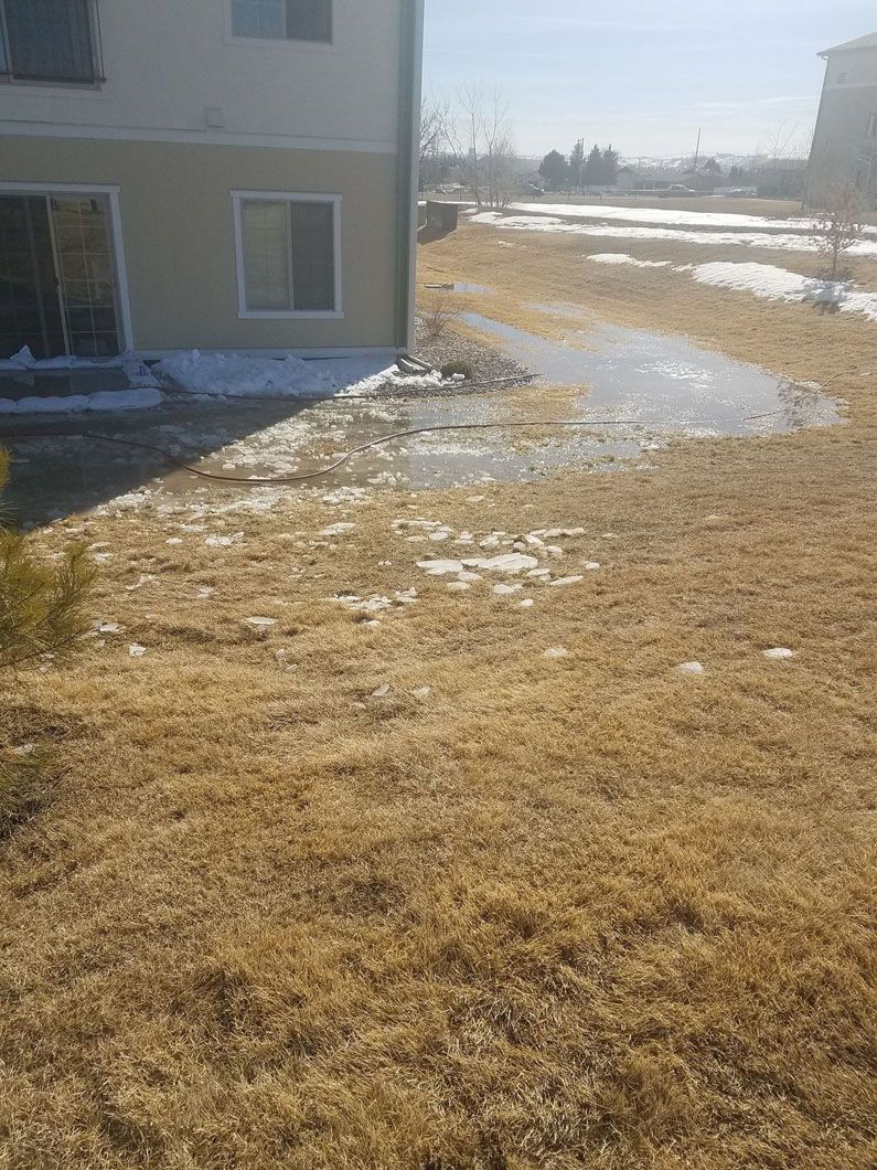 Snowy yard beside a tan house with patchy brown grass and a dirt path under bright sunlight