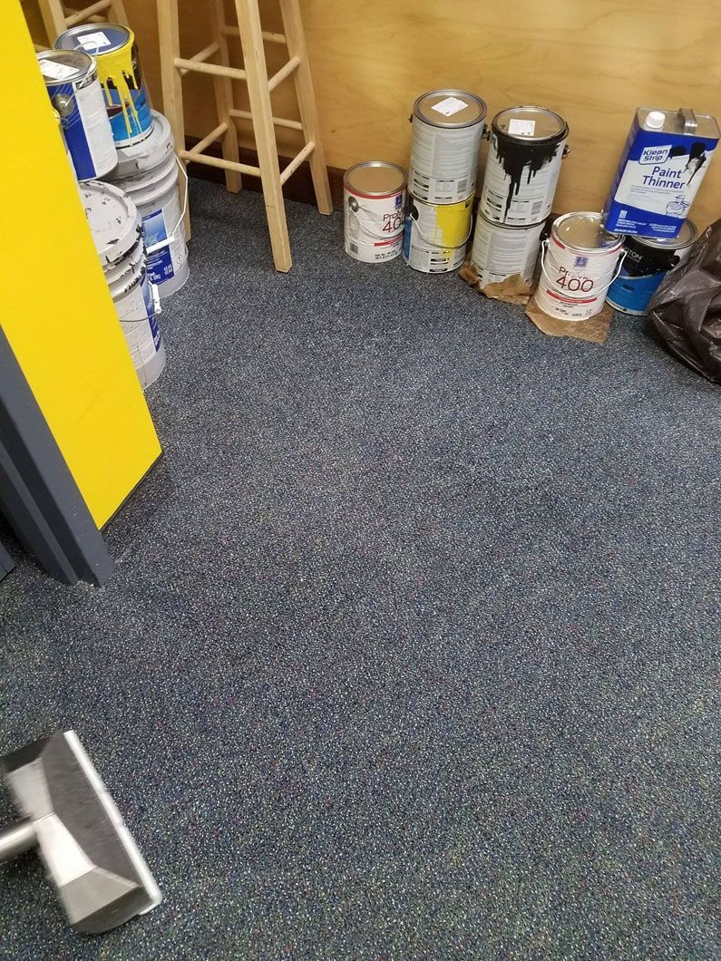 Paint cans and supplies stored on a gray carpeted floor beside a yellow wall and wooden shelf.
