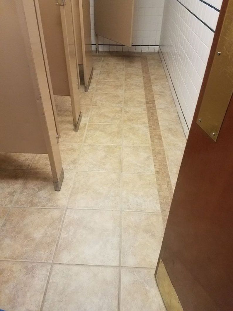 Narrow beige tiled hallway with restroom stall doors and a closed brown door at right.