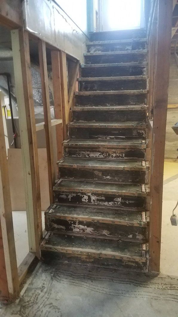 Steep worn wooden staircase inside an unfinished house, with exposed framing and a bright window above.