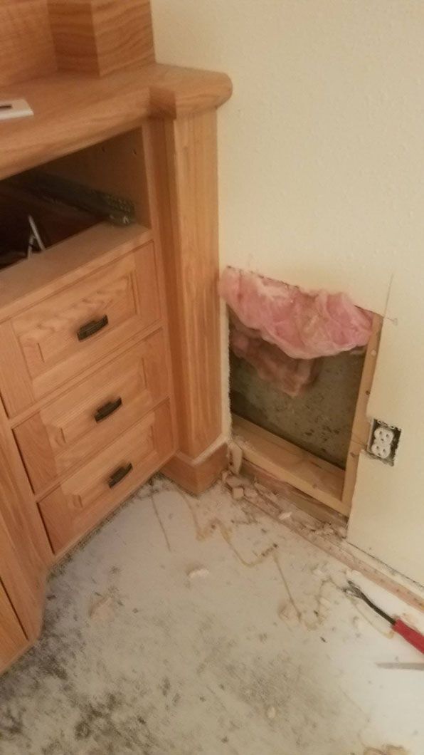 Wooden dresser beside a wall with a large damaged hole revealing pink insulation on the floor.