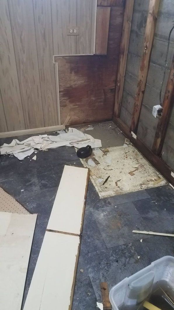 Damaged room corner with torn flooring, scattered debris, and exposed wall panels.