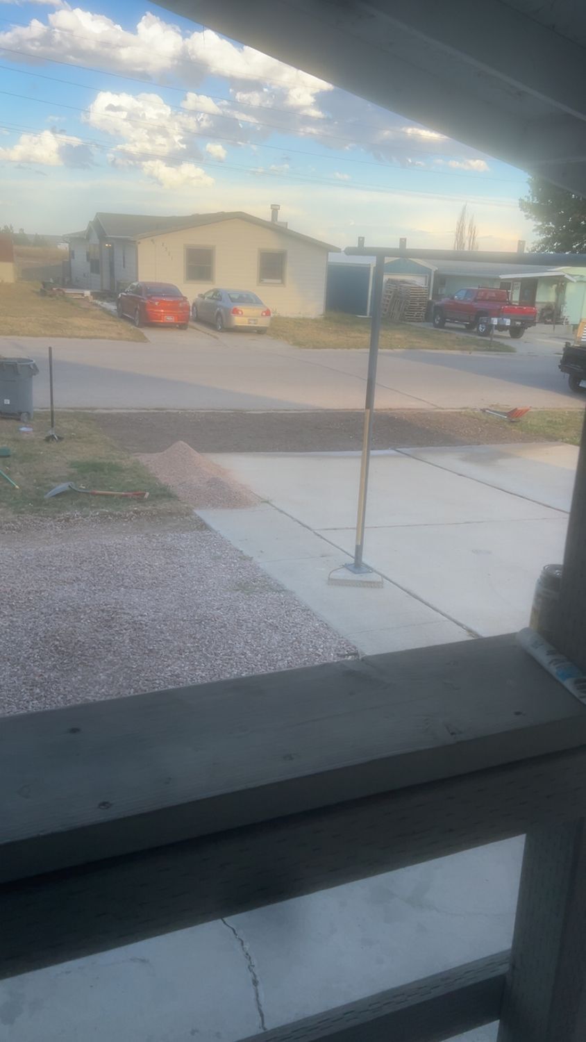 View through a window of a street with houses, parked cars, and a driveway under a cloudy sky.