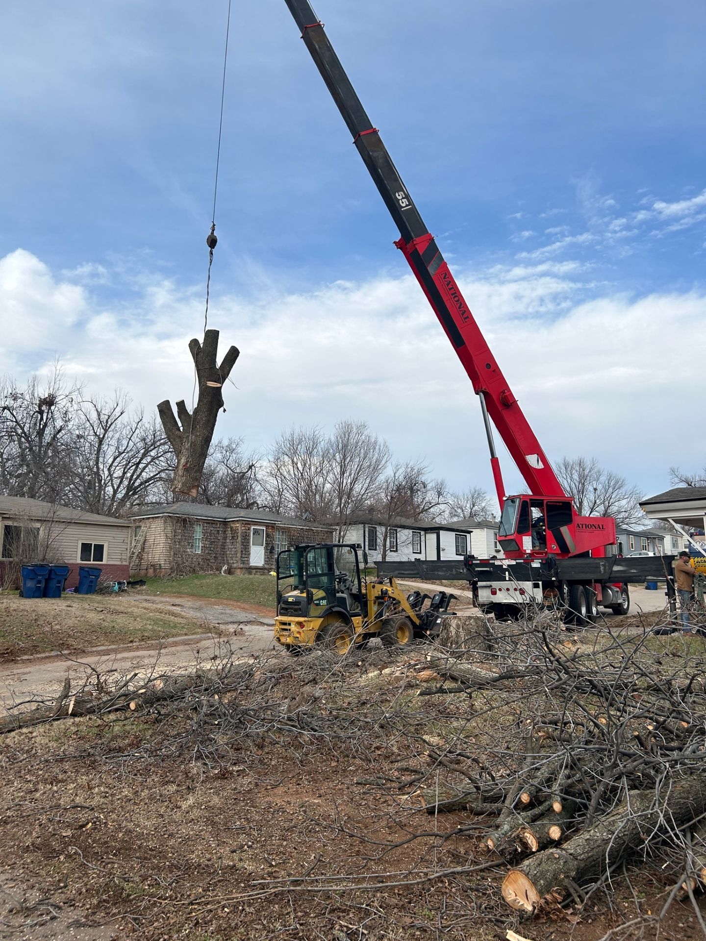 Crane lifting a large tree trunk at a worksite, with a red boom and yellow loader under a cloudy sky