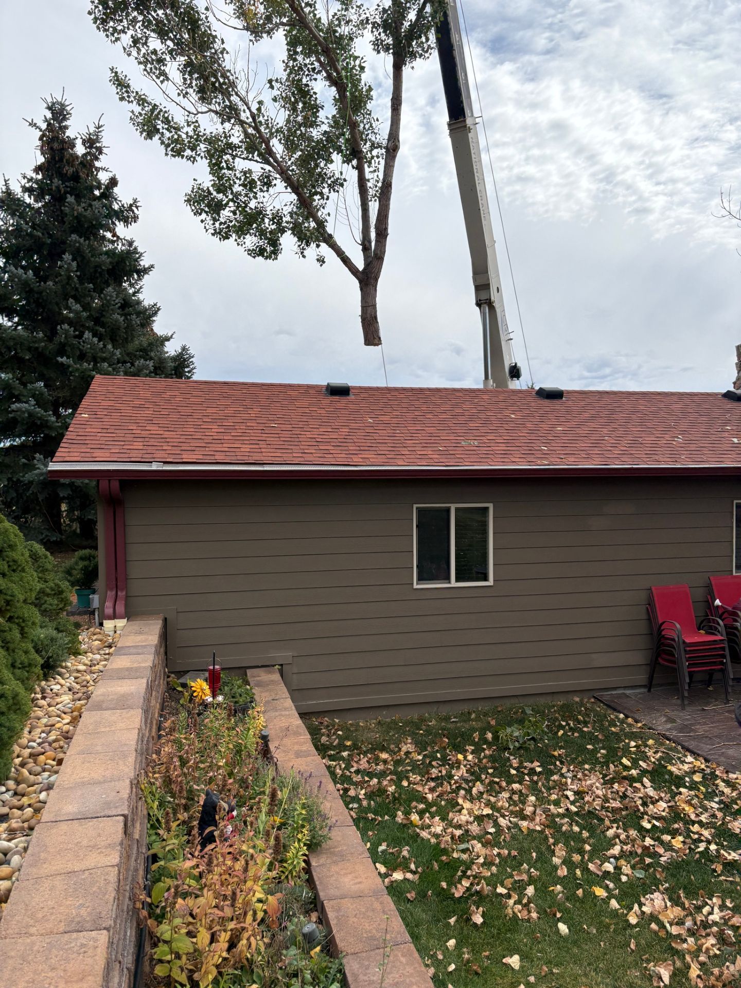 Backyard view of a brown house with a red roof, brick planter, and a crane lifting a tree overhead