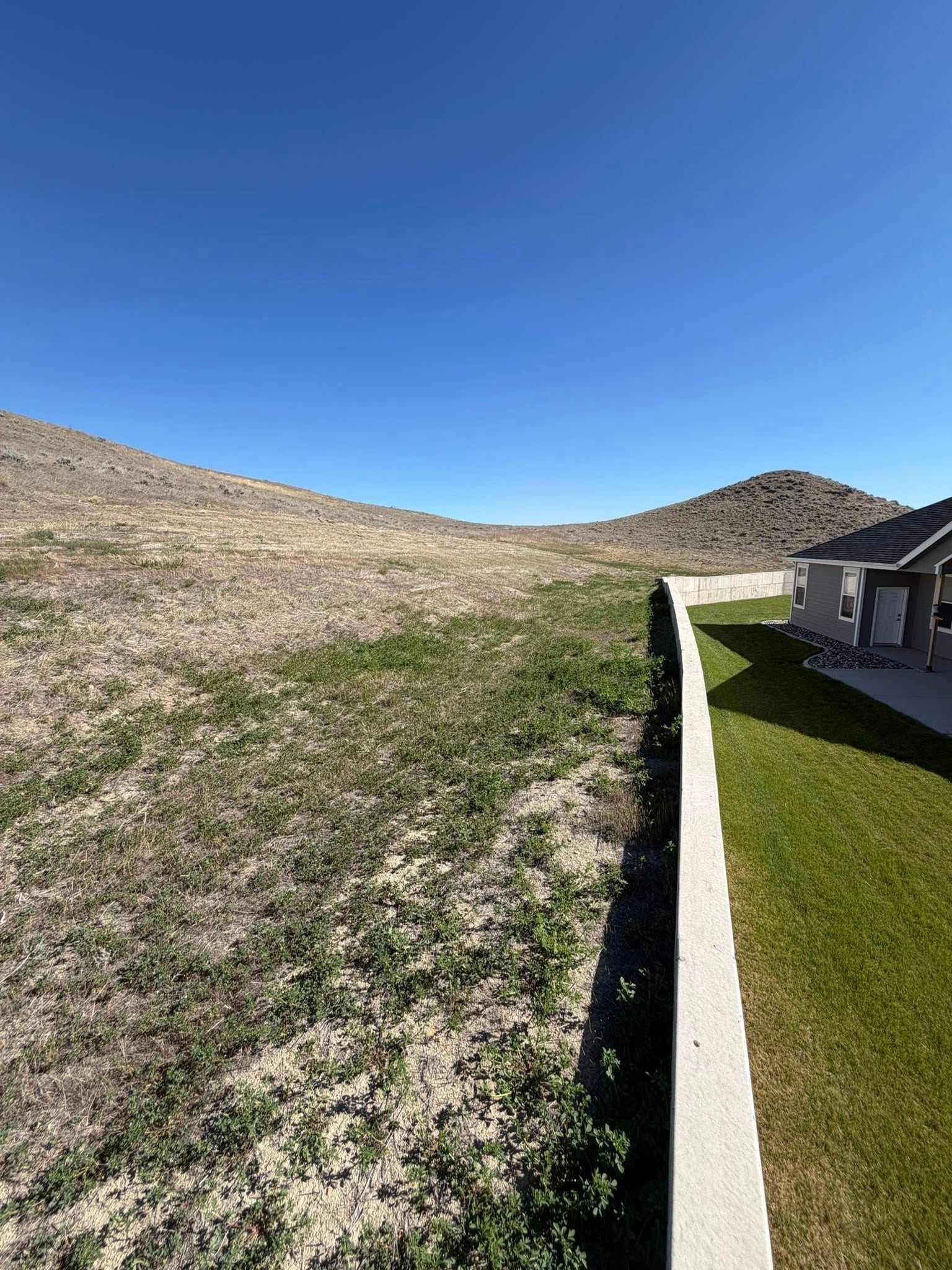 Desert hillside beside a manicured green lawn and white wall under a clear blue sky.