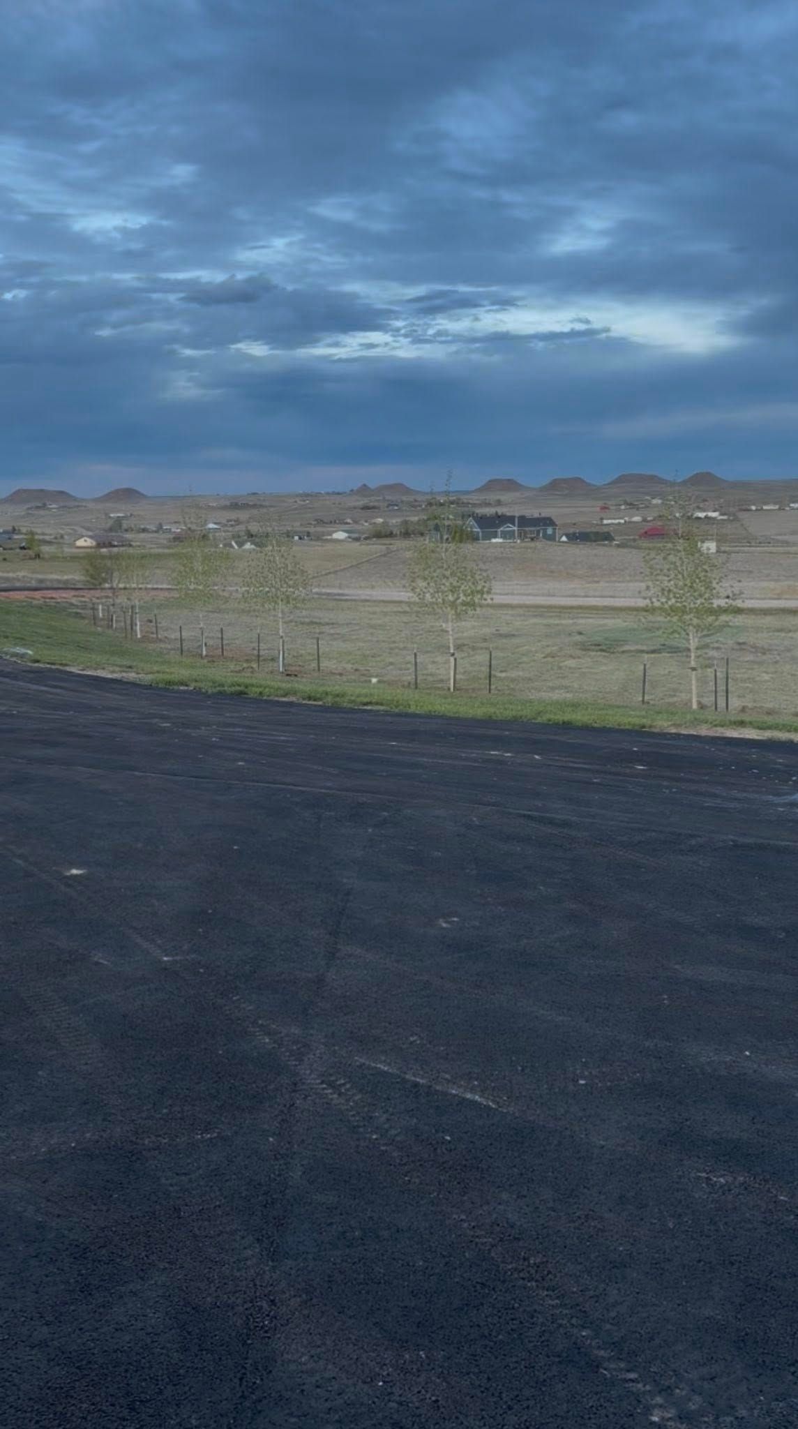 Dark asphalt path beside a grassy field under a cloudy blue sky, with houses in the distance
