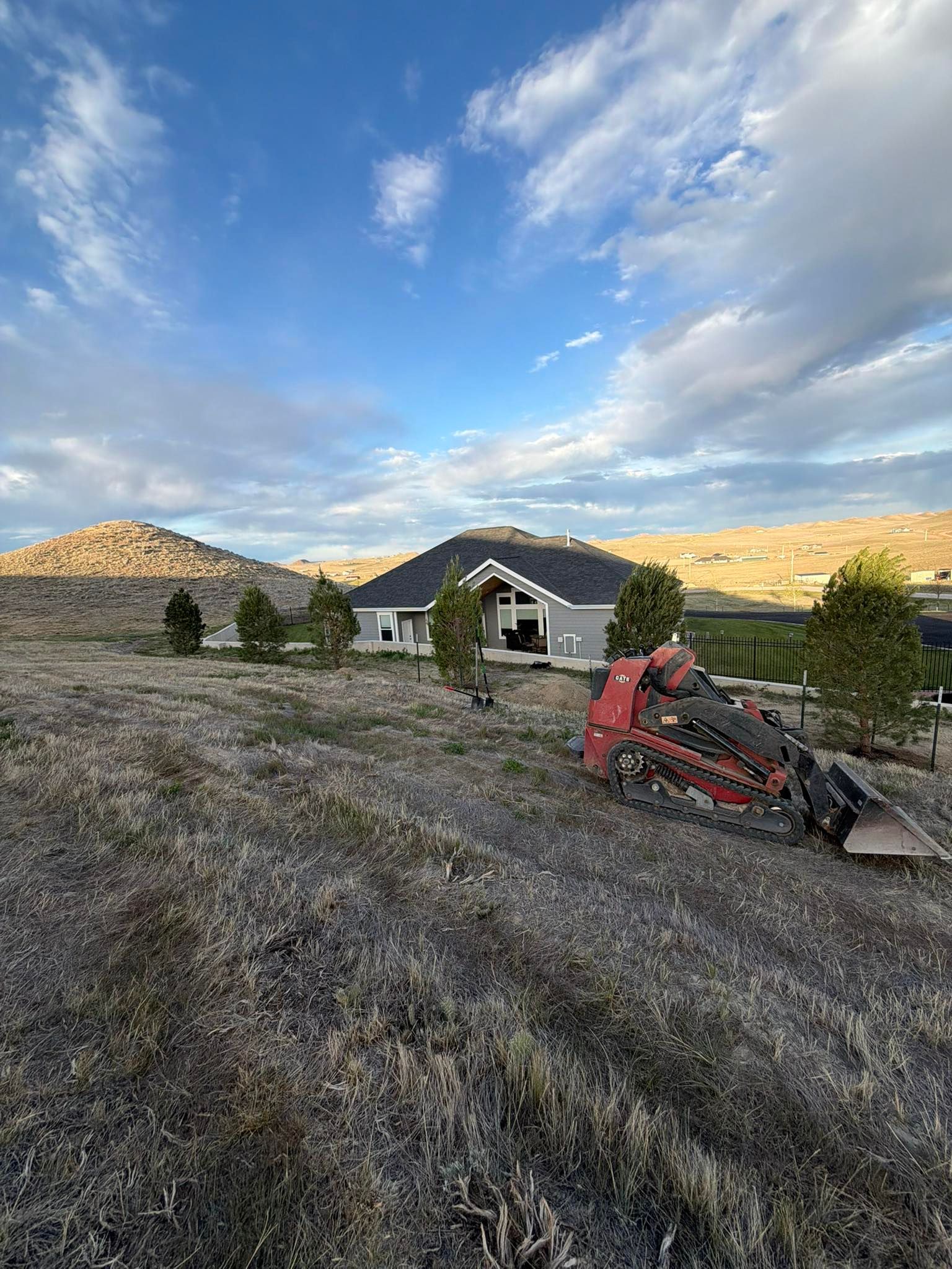 Person operating a red mower on a dry hillside with a white house, mountains, and cloudy sky in the background