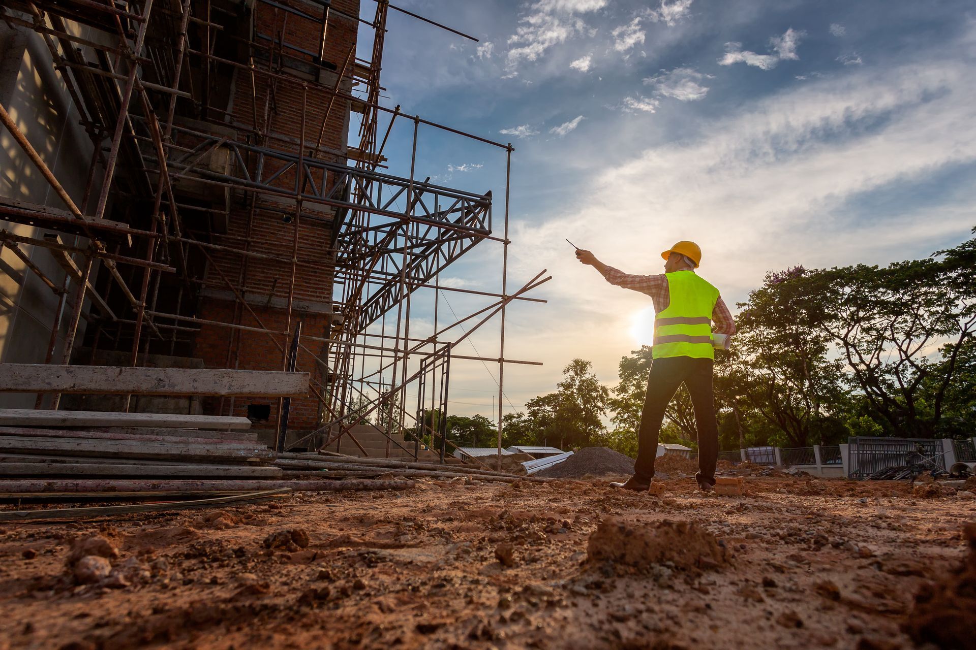 Construction worker in neon vest directing on a muddy site beside scaffolding at sunset