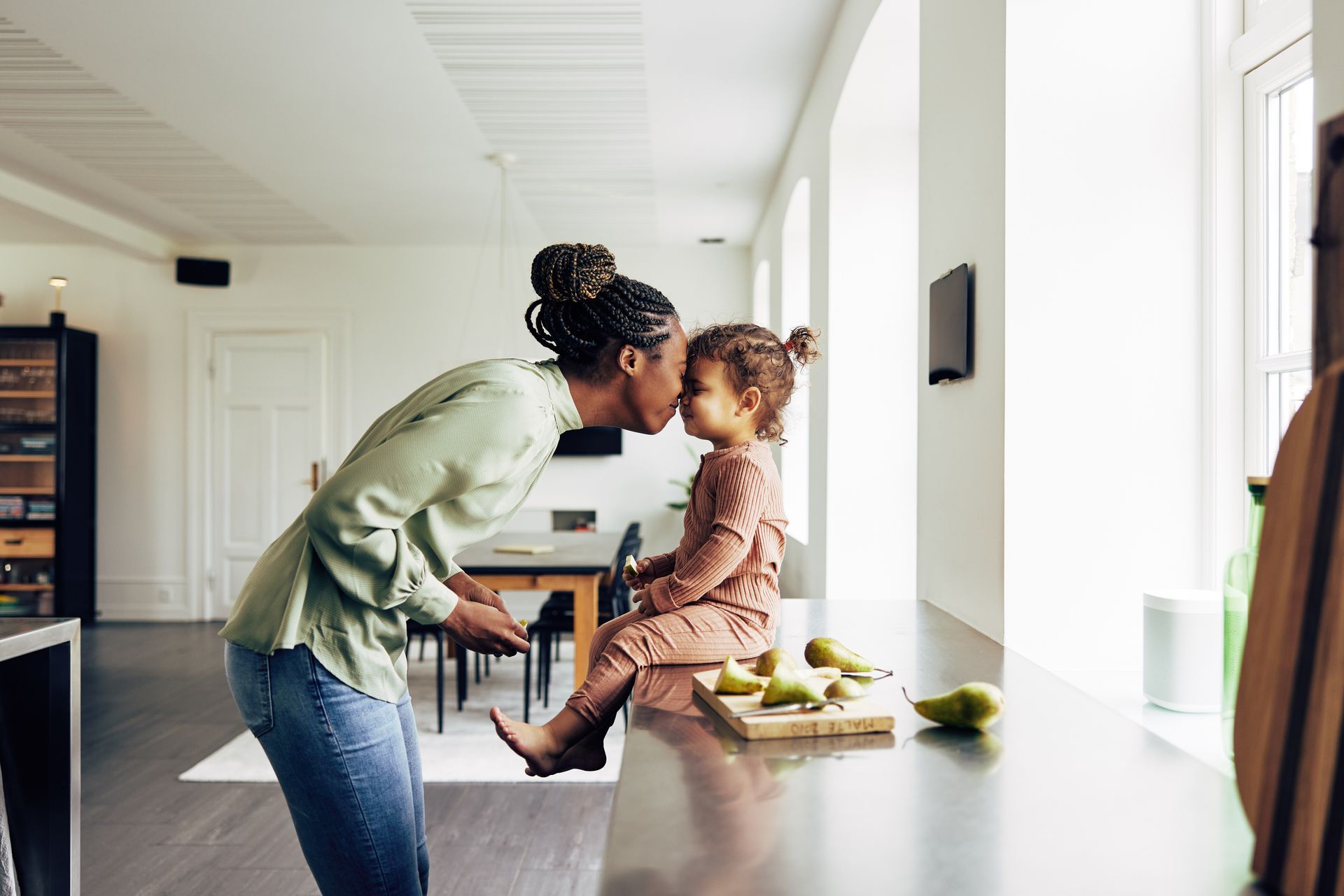 Woman kissing child in a bright kitchen beside a table with fruit