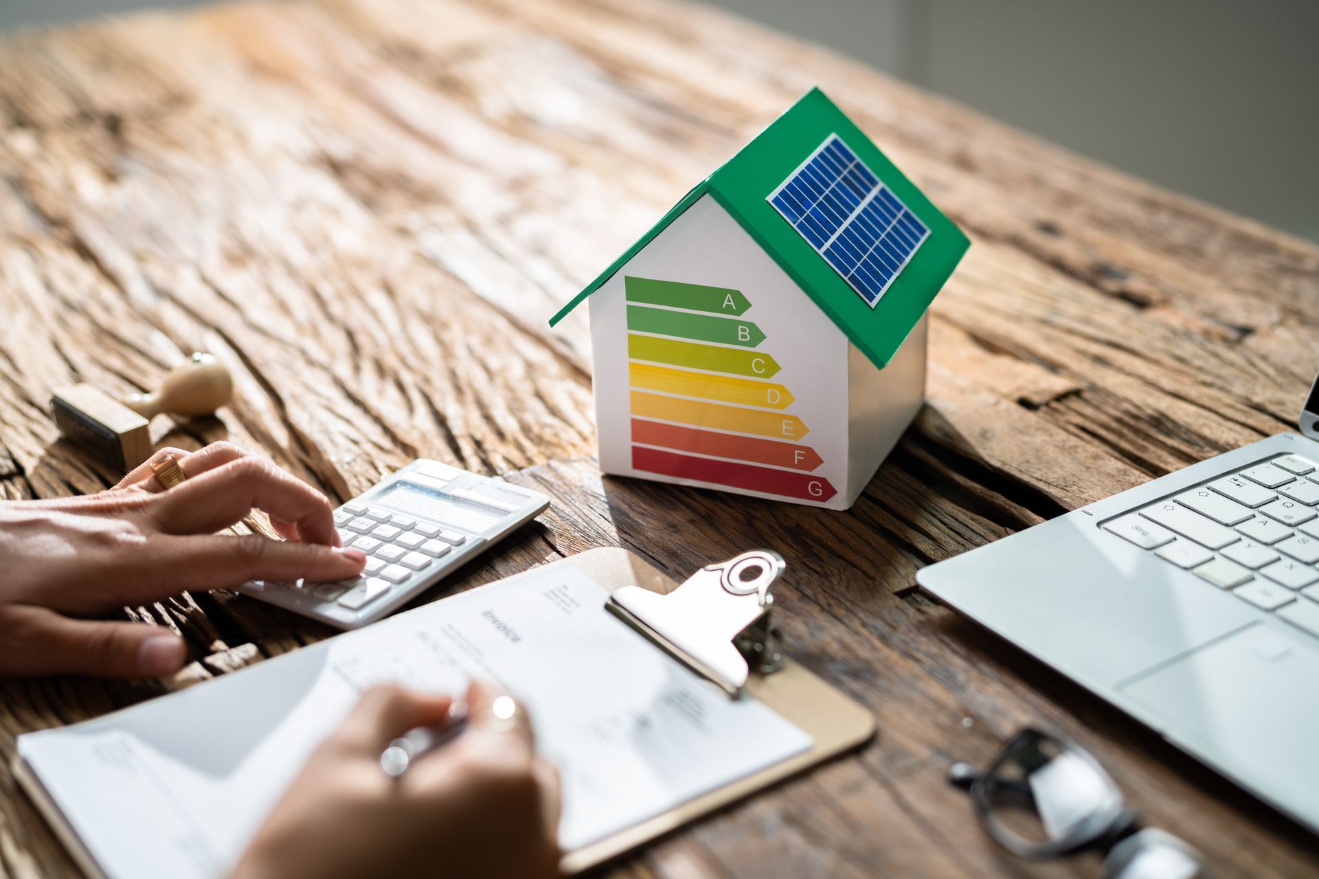 Hands using a calculator beside a house model with green roof and energy rating chart on a desk
