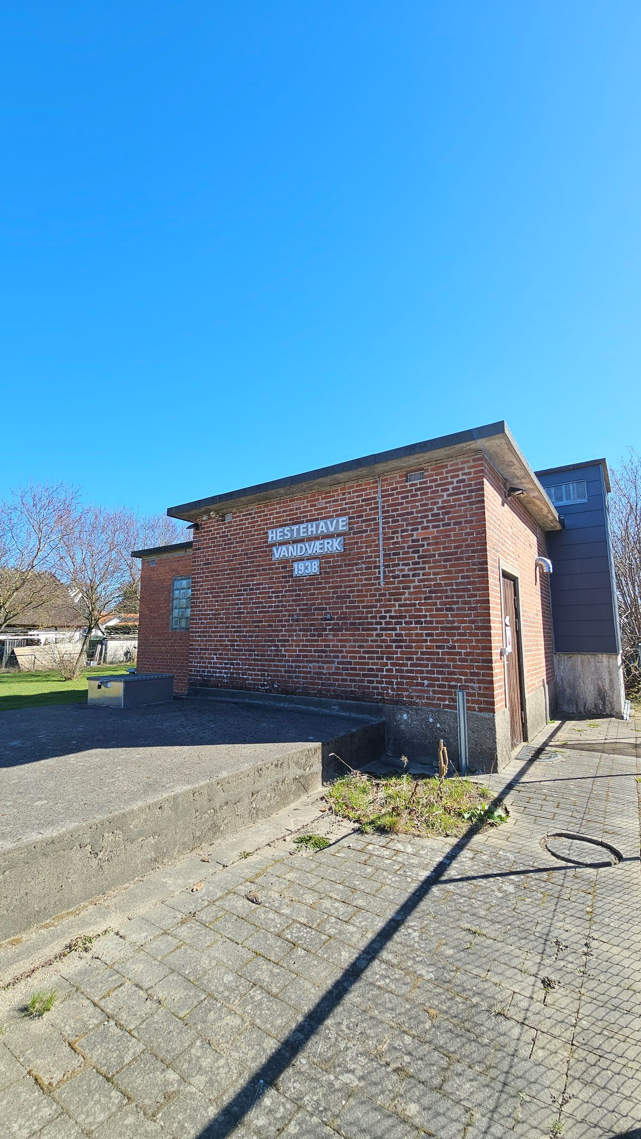 Small brick building with a mural, under a bright blue sky, beside a paved area and a tree in bloom