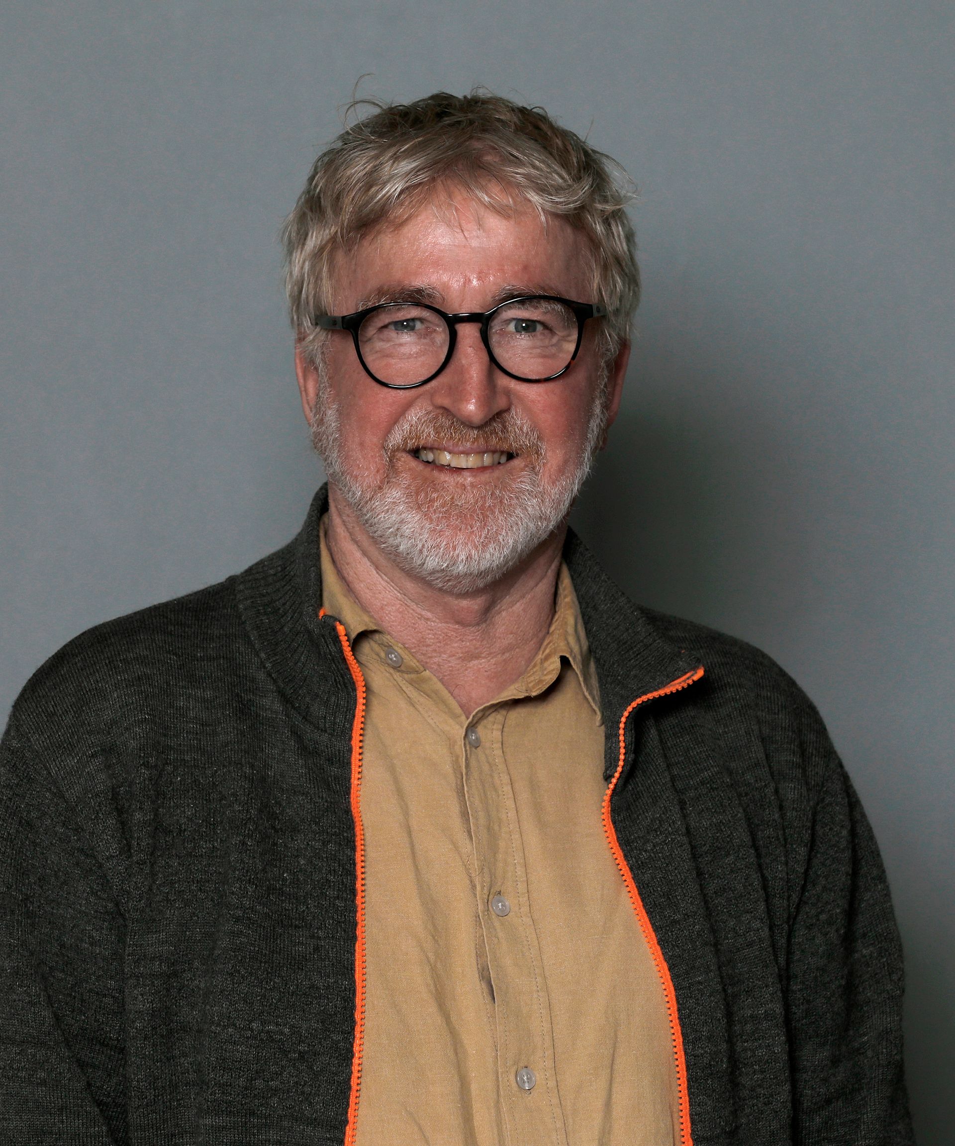 Smiling man with glasses and gray beard wearing a tan shirt and dark jacket against a gray background