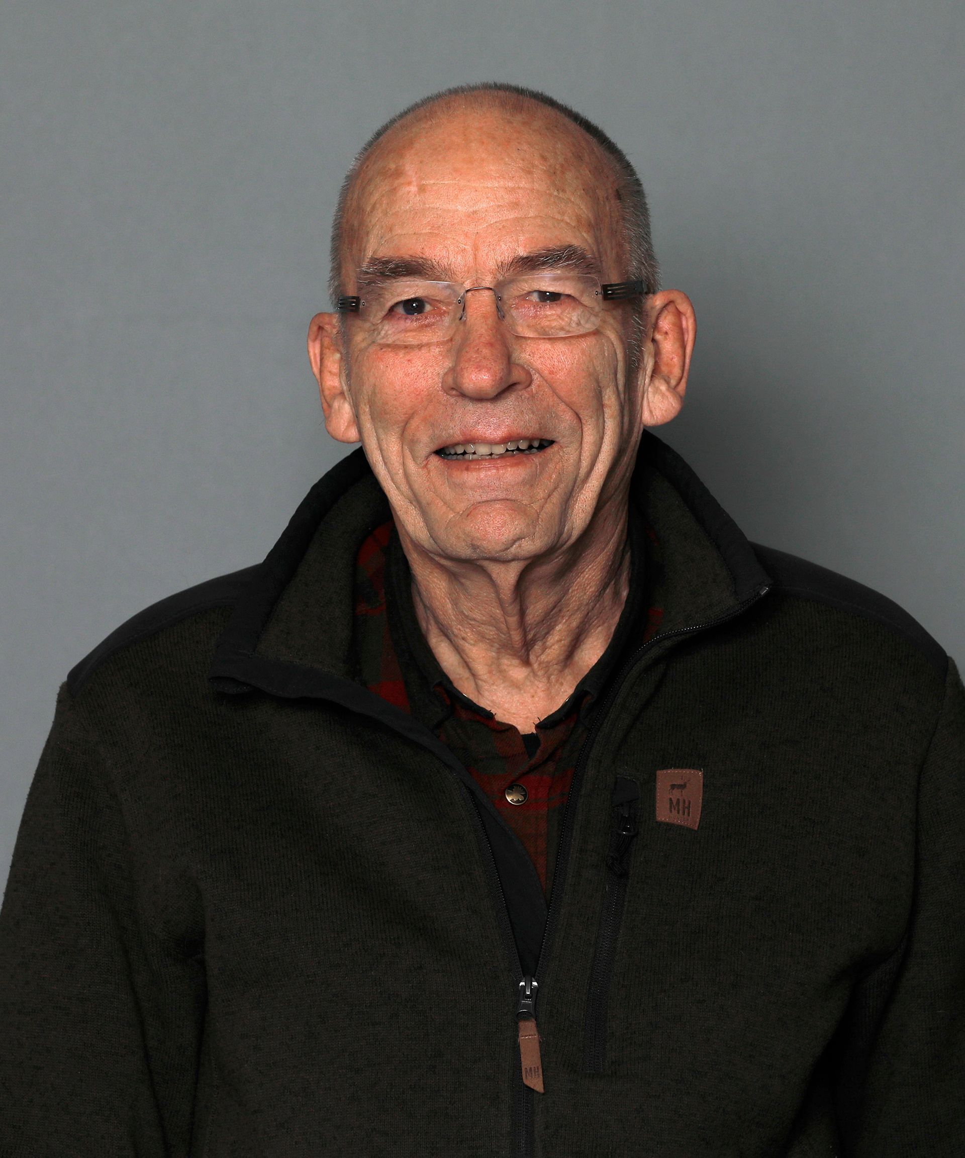 Smiling man in glasses wearing a dark jacket against a plain gray background