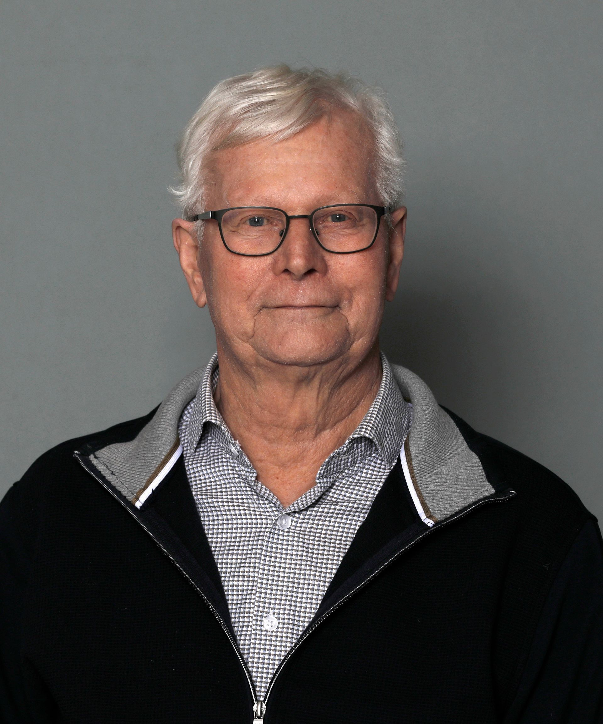 Portrait of a man with glasses wearing a black jacket over a checkered shirt against a gray background