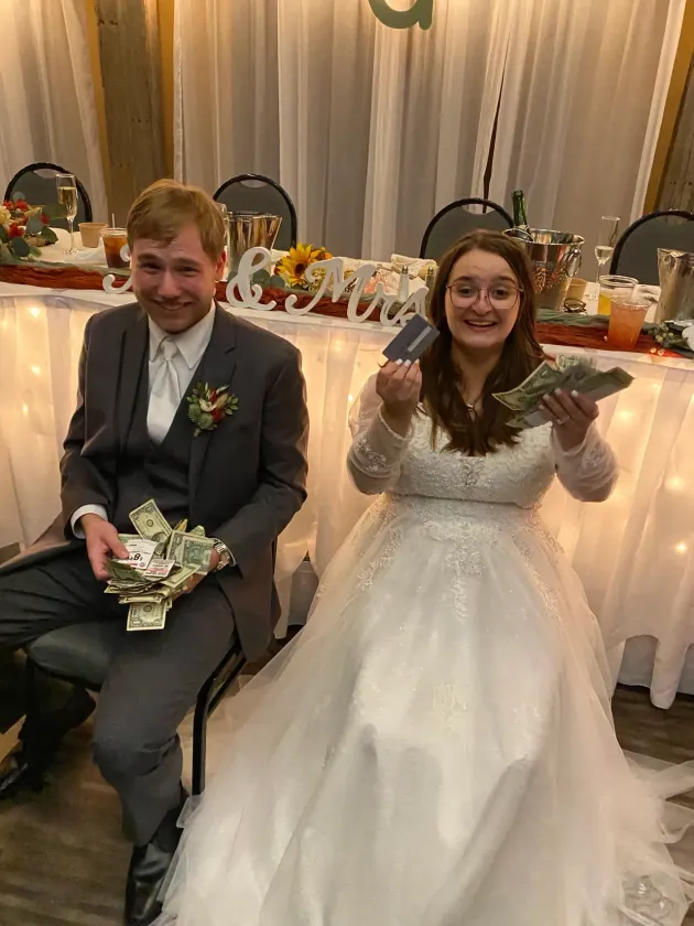 Newlyweds seated, holding cash and a card at their wedding reception.
