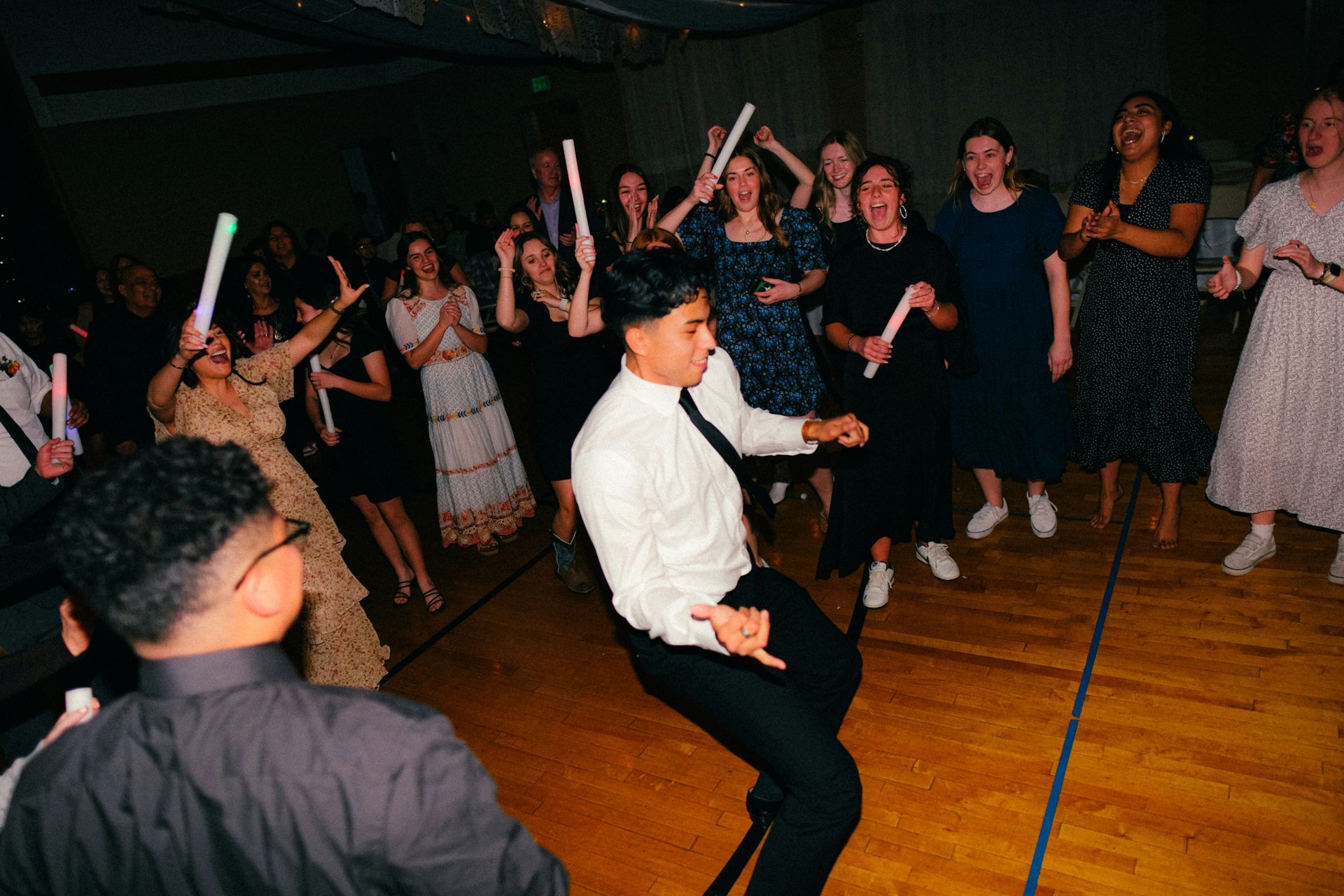 People dancing in a well-lit room, some holding glow sticks. One man is doing a dance move.