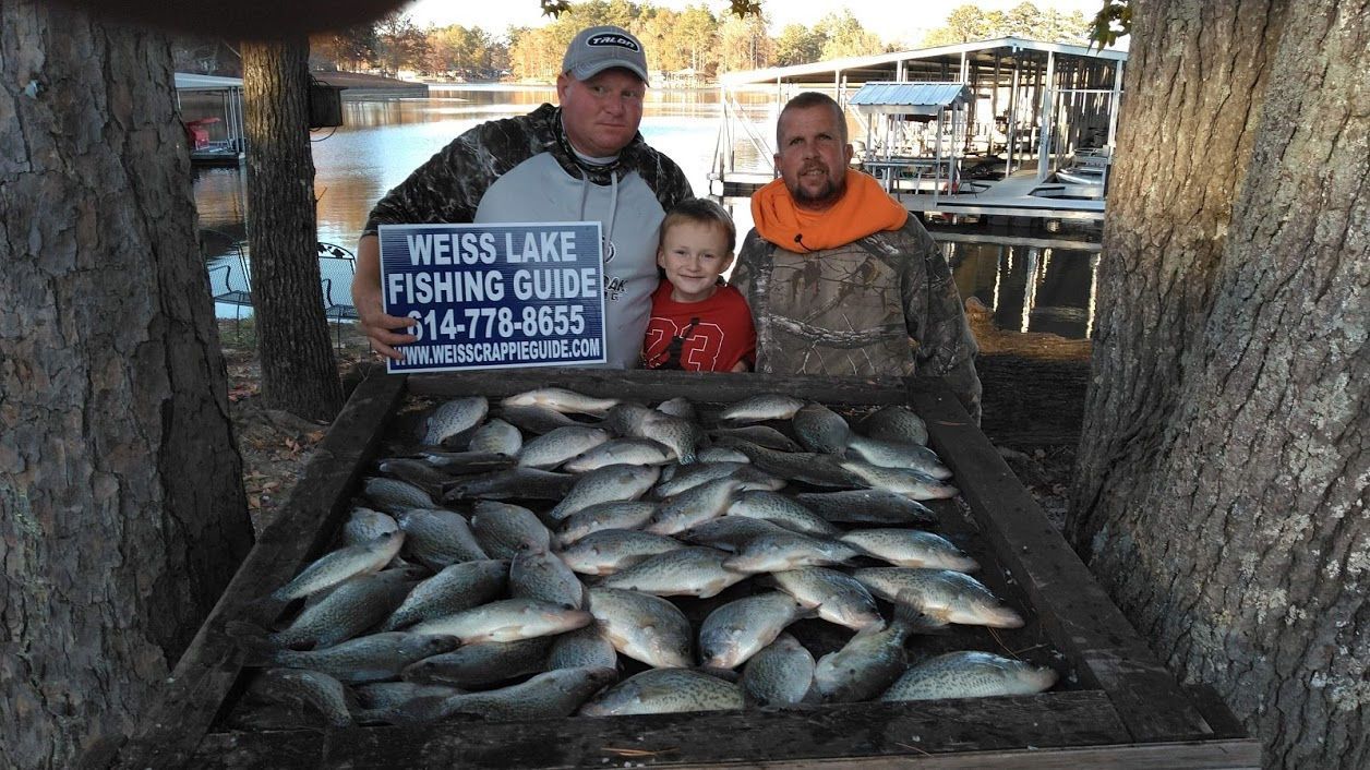 Two people and a child stand behind a wooden crate filled with a large catch of fish, holding a Weiss Lake fishing sign.