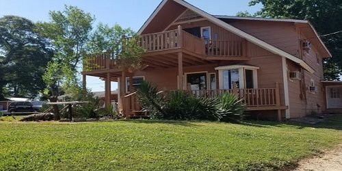 A two-story, light-colored wooden house with a spacious deck and balcony, set on a green lawn under a clear blue sky.