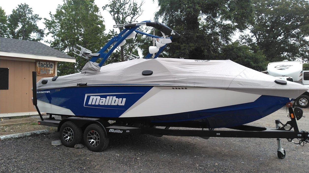 A blue and white Malibu ski boat on a trailer, covered with a grey tarp, parked outside a building.
