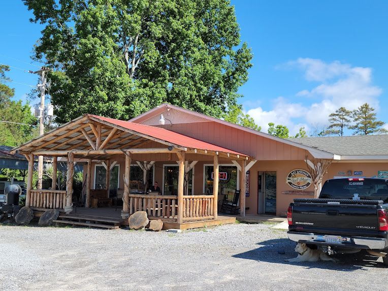 A rustic building with a wooden porch and red metal roof, featuring a gravel parking lot and a parked black pickup truck.