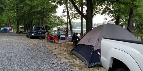 A campsite near a lake with tents, a parked SUV and pickup truck, and a group of people sitting in lawn chairs.