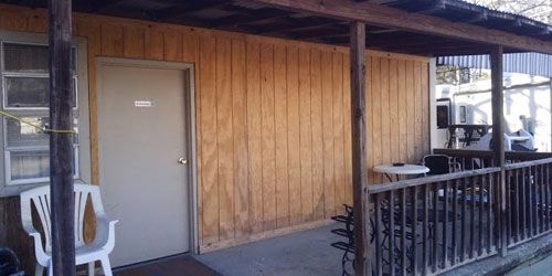 A wooden cabin with a beige door, a window, a white plastic chair, and a covered porch area with a small table.
