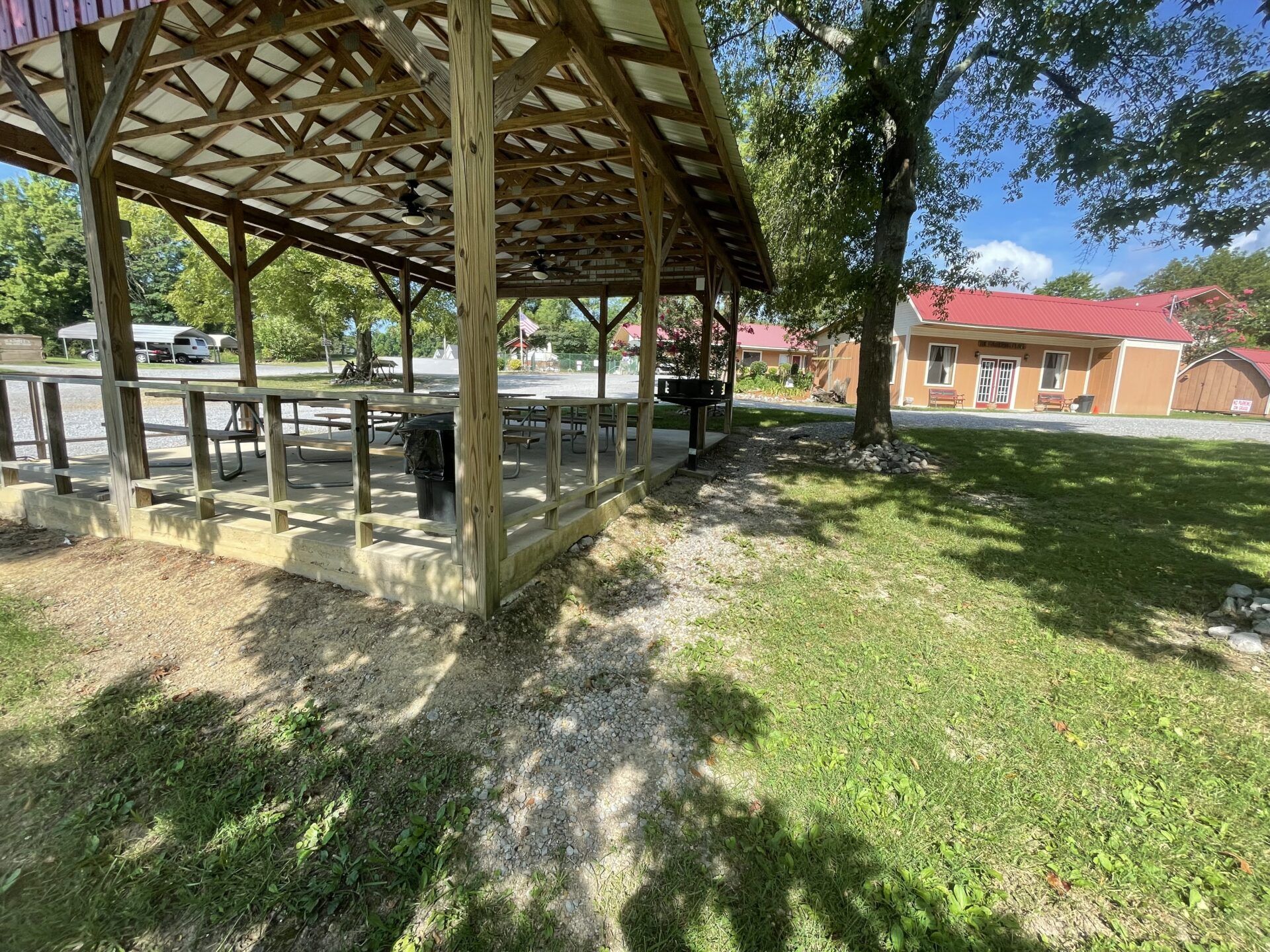 A wooden pavilion with a metal roof stands over picnic tables on a sunny day next to a grassy lawn and a tan building.