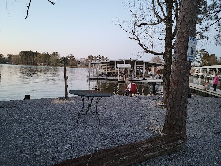 A round metal table sits on a gravel shore overlooking a calm lake with a covered boat dock in the distance at sunset.