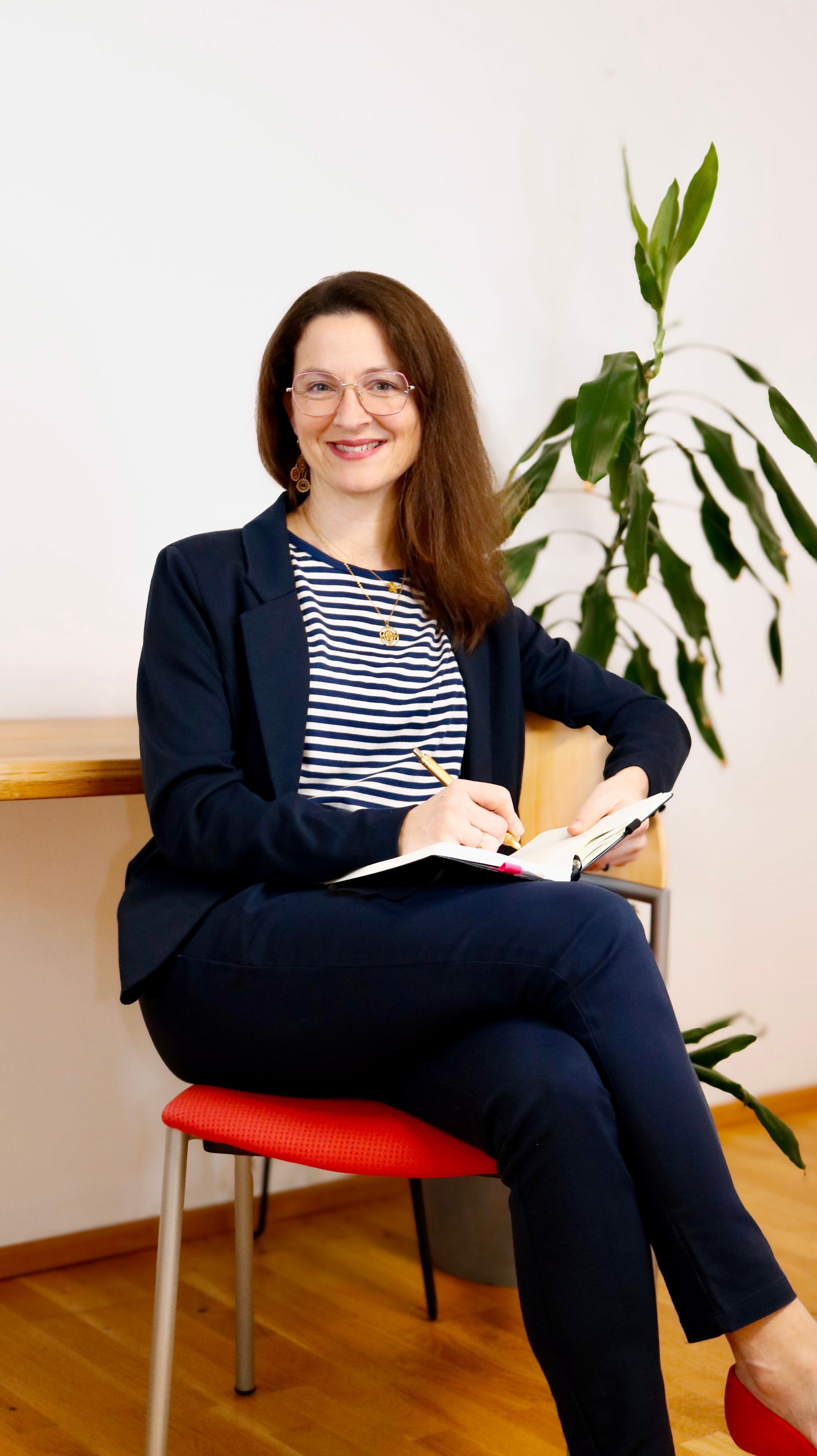 Eine lächelnde Person mit langen Haaren, beigem Oberteil und heller Hose sitzt in einem türkisfarbenen Sessel vor einer schlichten Wand.